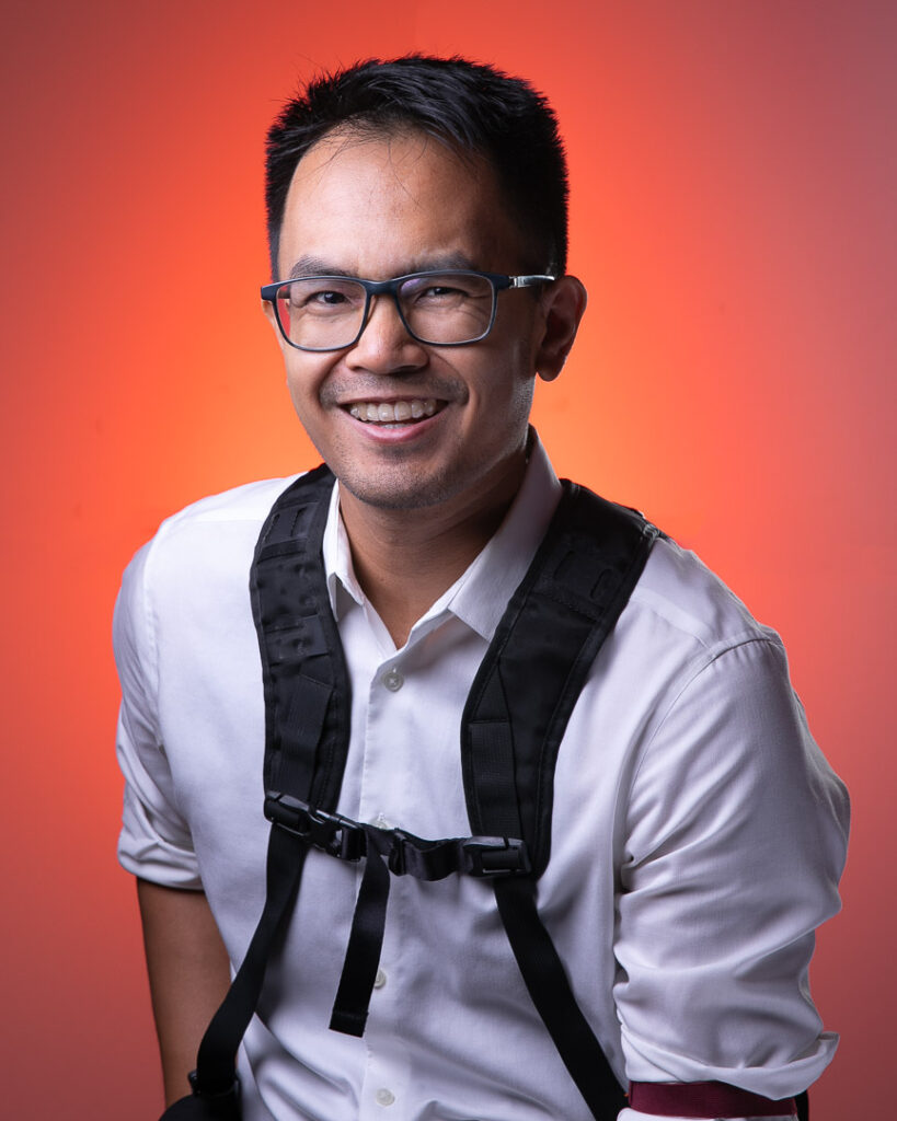 Edmar Castillo wearing black-framed glasses while wearing white dress shirt and dual camera straps posing and smiling in front of red backdrop 